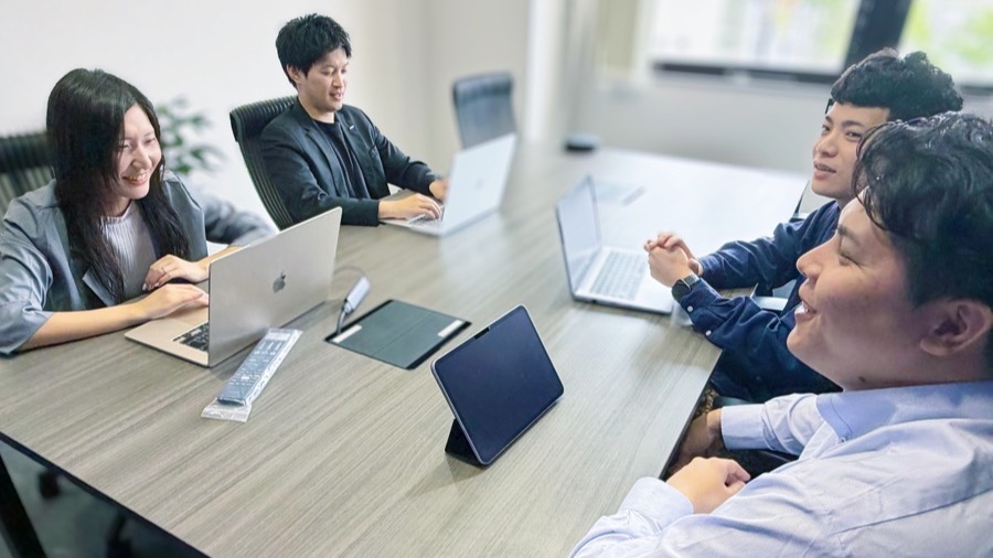 会議テーブルを囲んでノートPCを開き、笑いながら話すメンバー